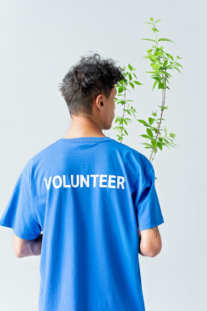 Person in blue volunteer shirt carrying a green plant, viewed from behind.